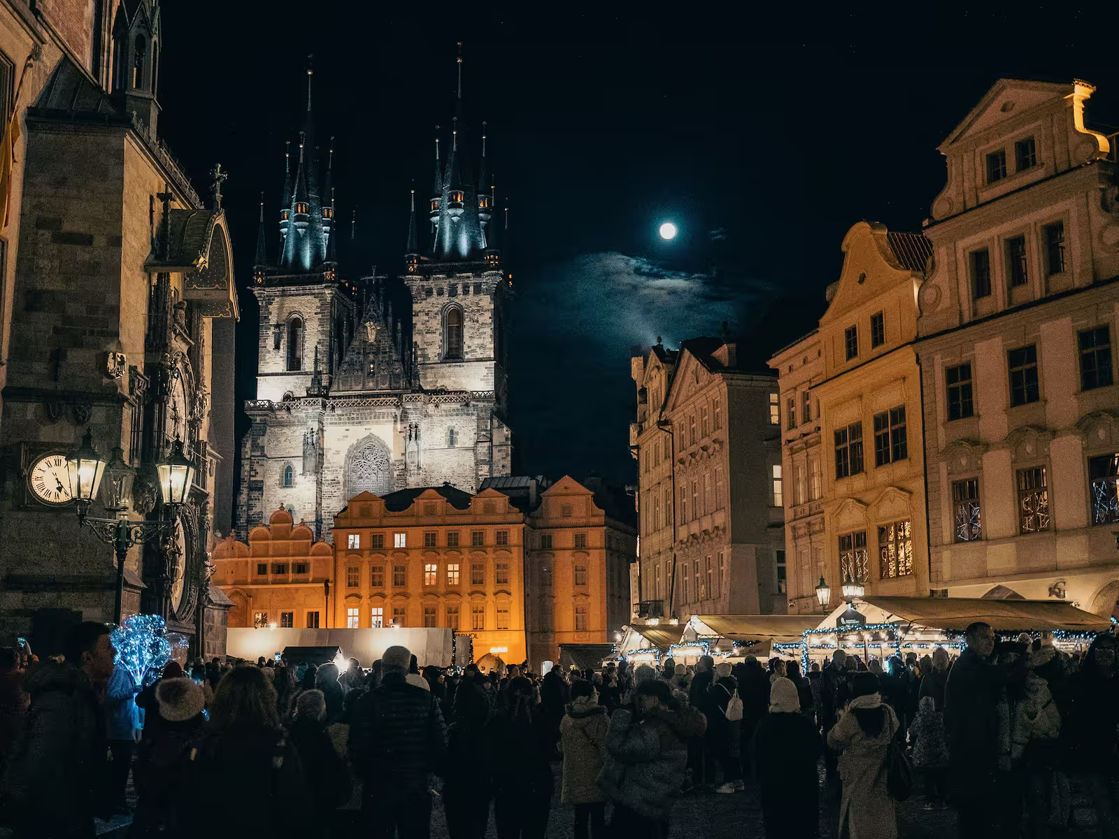 Groups of people in Old Town Square Prague at night with moon in sky and Christmas markets