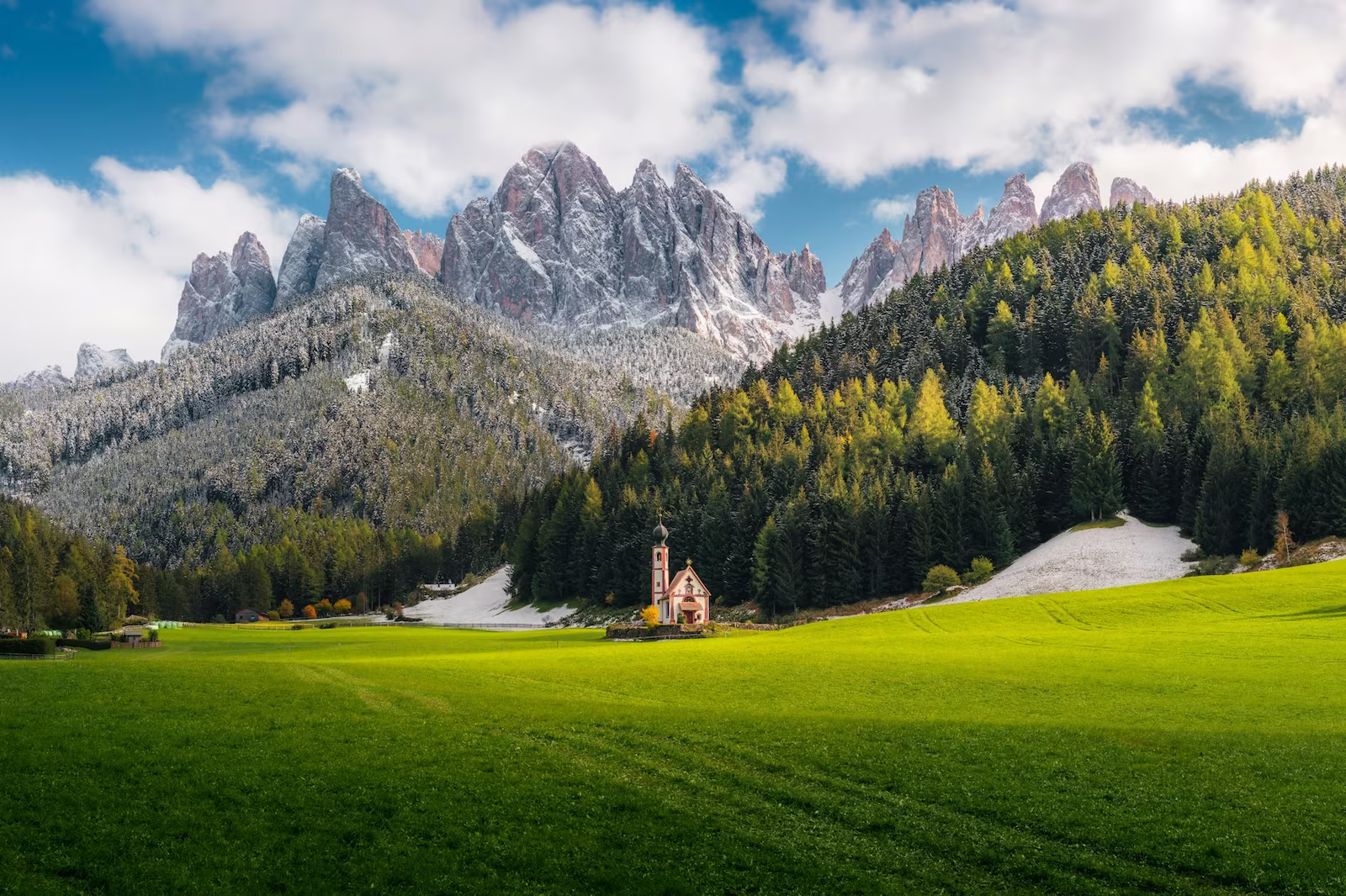 small church on a green grassy plain with towering tree and snow filled mountains in the background