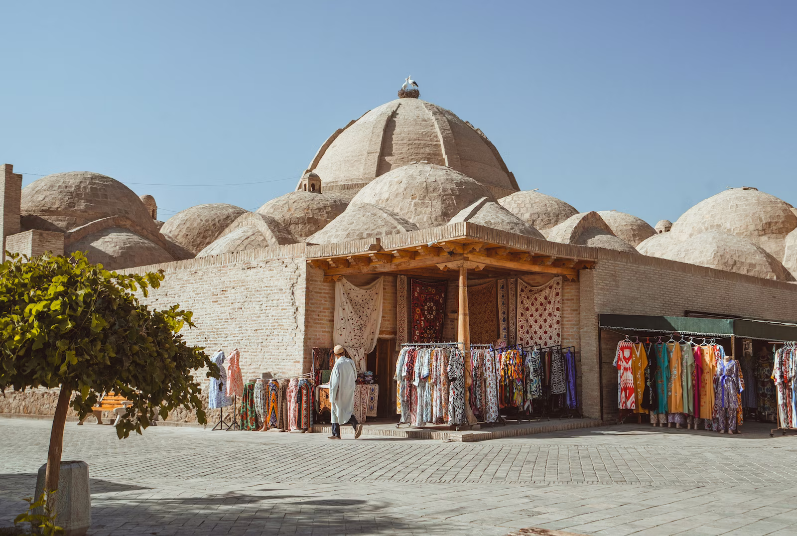 man in white robes walking past a market selling colourful textiles