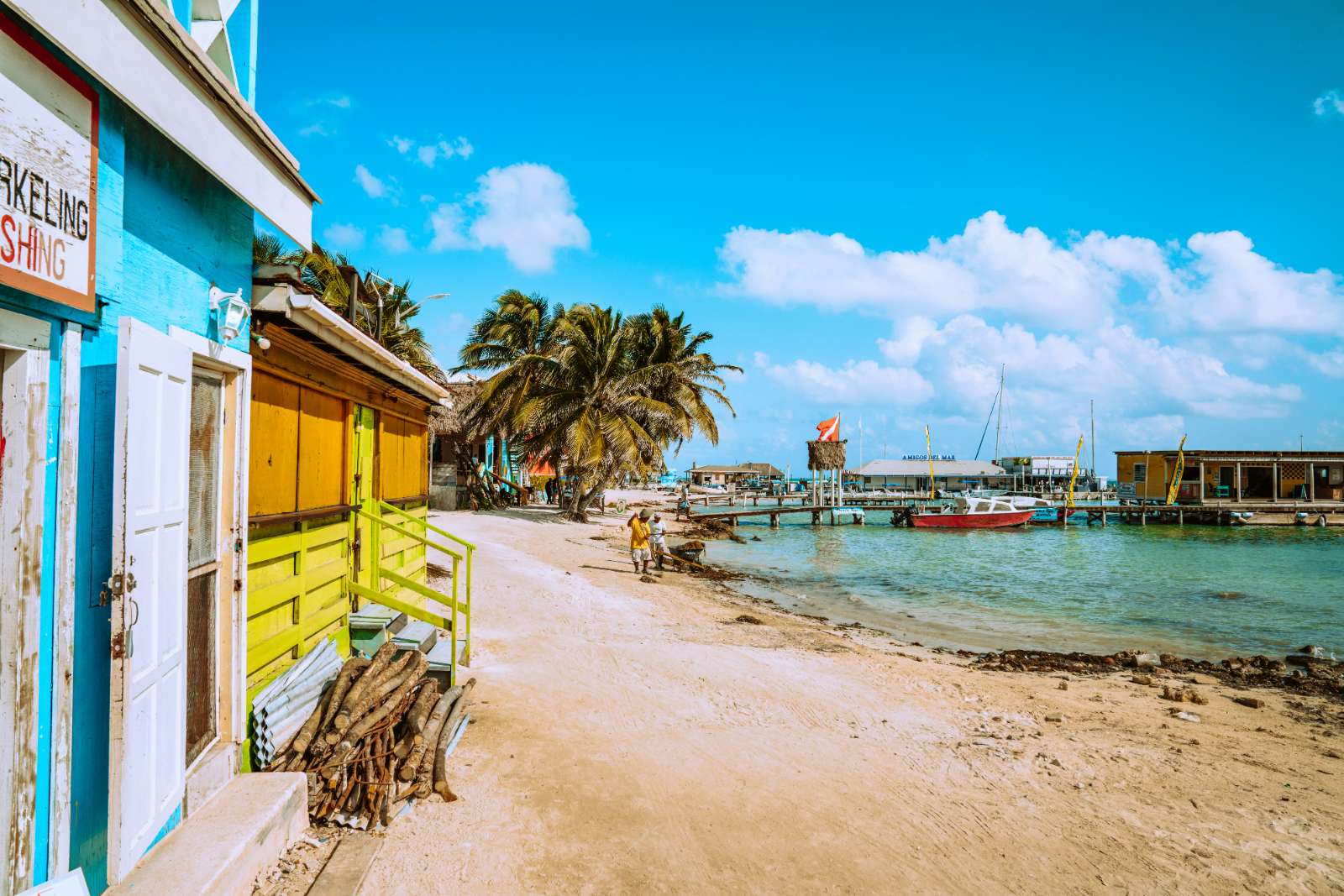 Bright beach huts on sand banks of bear with pier, boats and palm trees in background