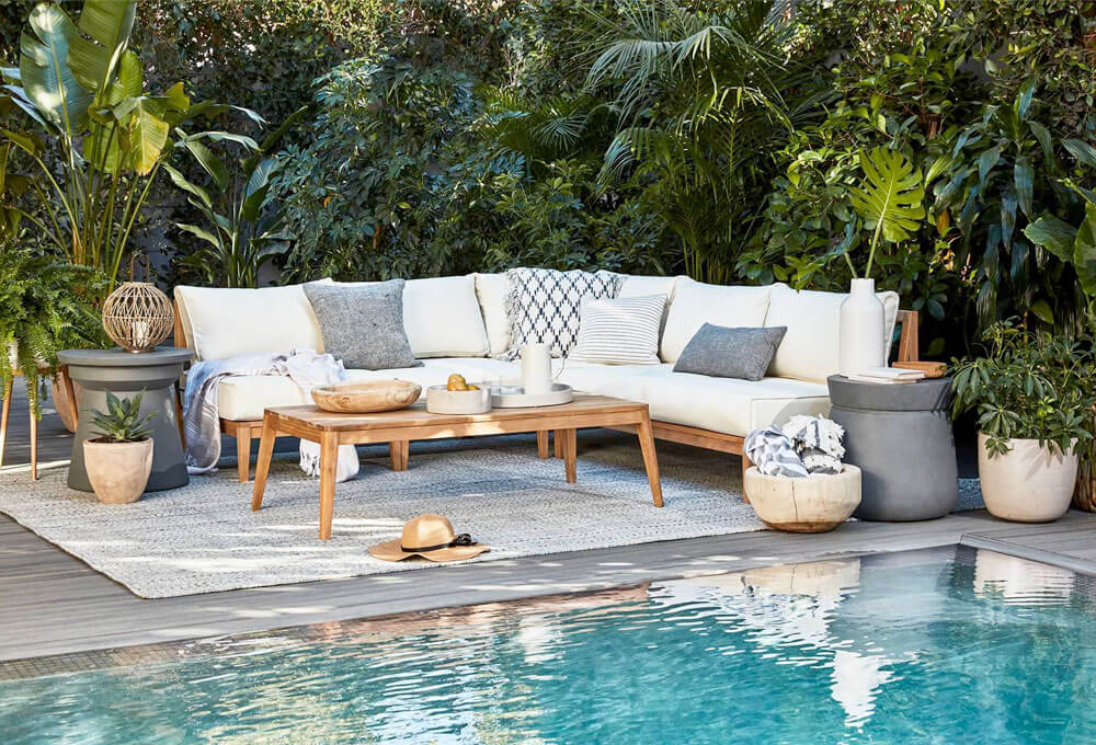 Teak Outdoor Sectional with white cushions, blankets and a wooden coffee table near plants and pool