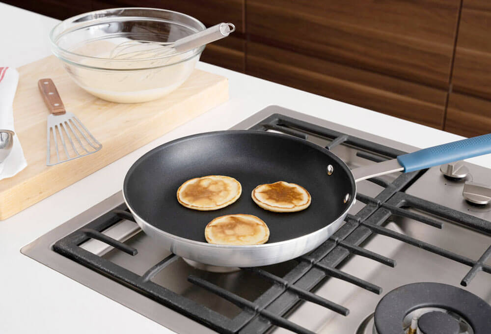Non-stick frying pan with blue handle cooks, three small pancakes on a gas stovetop