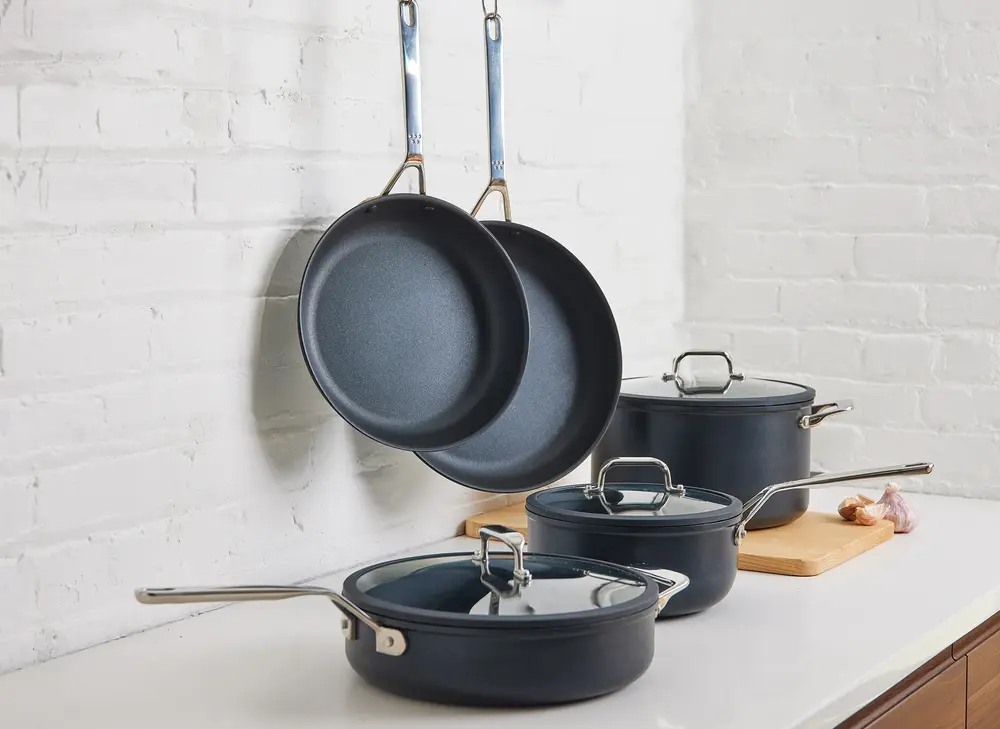 A set of dark nonstick pots and pans with glass lids, two of them hanging on a white brick wall