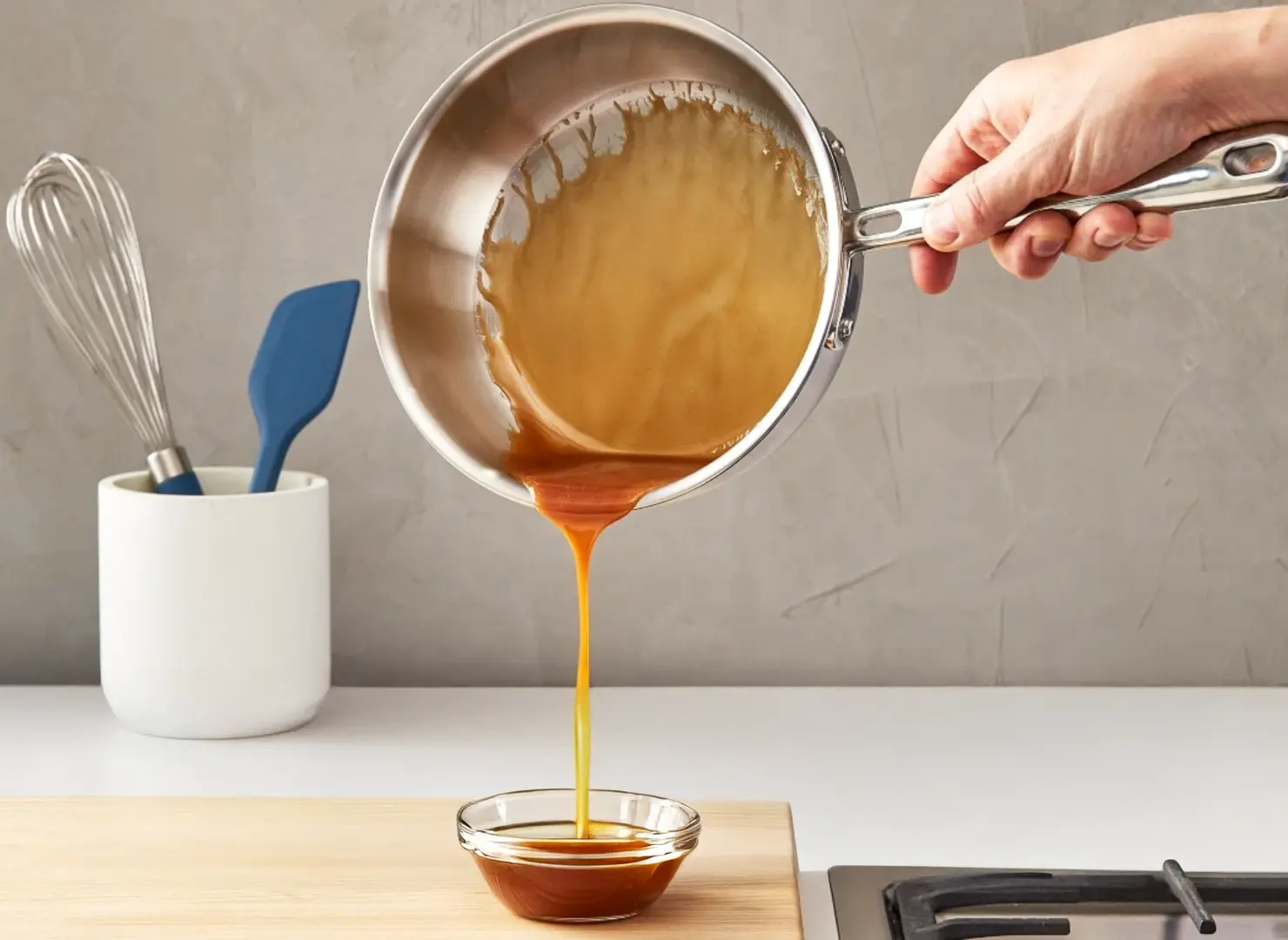 A person is pouring caramel sauce from a stainless steel saucepan into a small glass bowl 