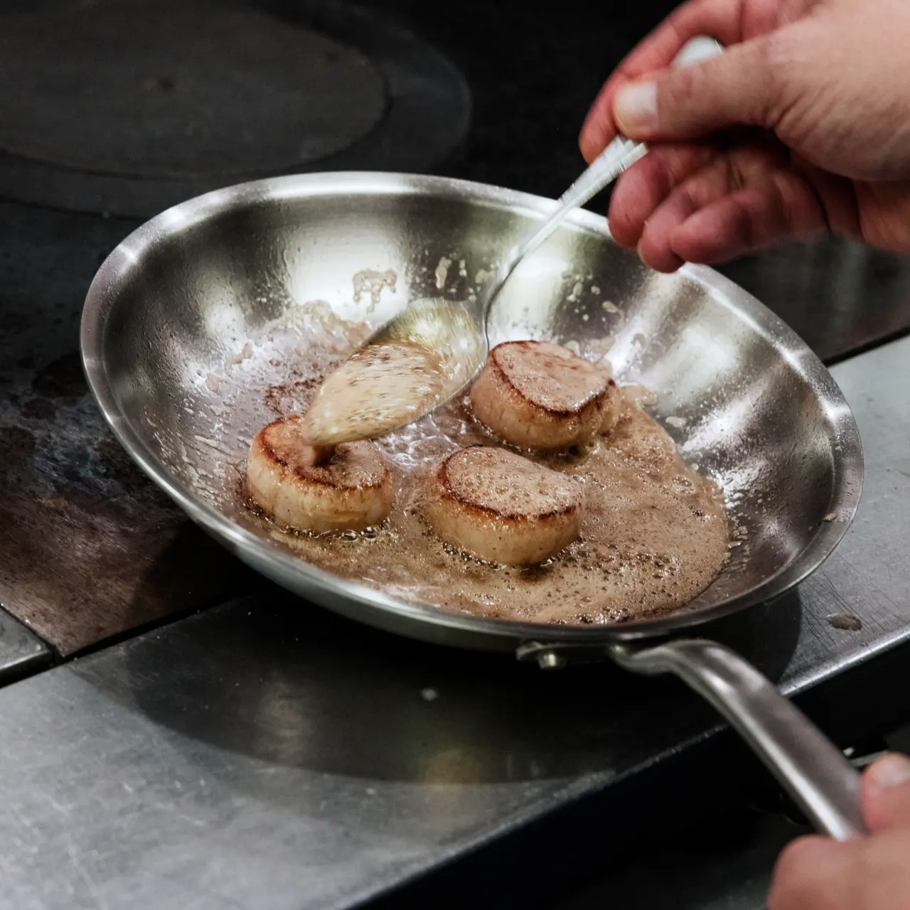 A person is basting seared scallops with butter in a stainless steel pan on a stovetop.