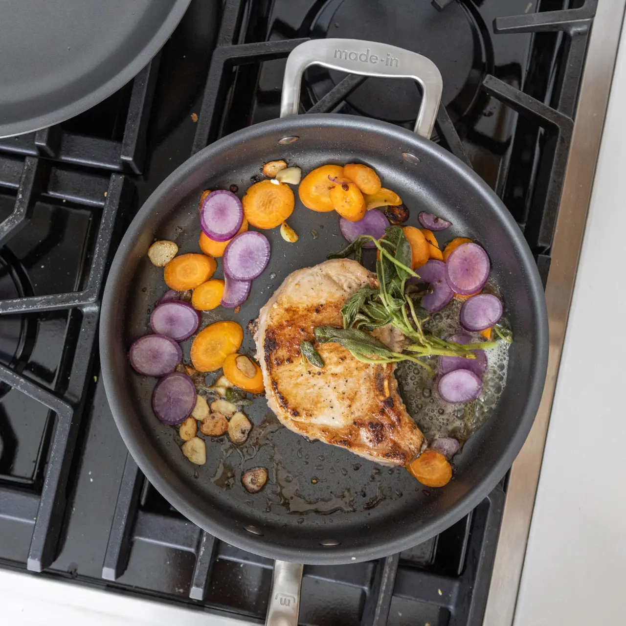 Nonstick pan n a stovetop holds a seared pork chop, sliced garlic purple and orange carrot and herbs