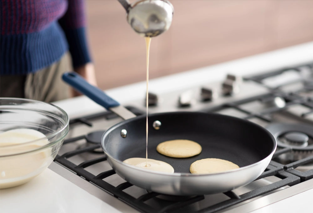 batter is being poured into a non-stick frying pan on a stovetop to make pancakes