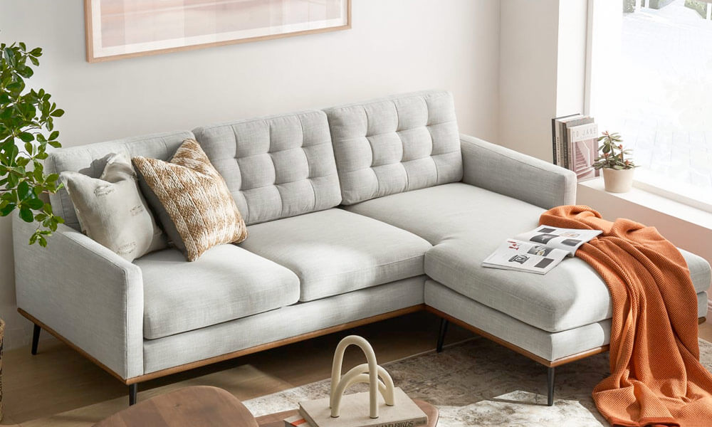 Grey upholstered sectional with grey and mustard throw pillows, an ottoman and an orange blanket