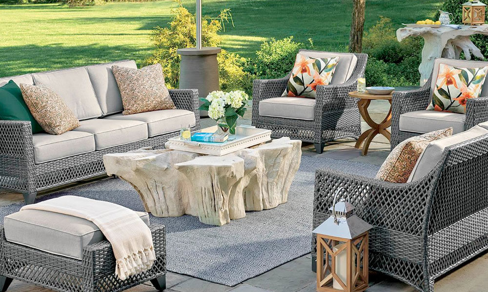 A patio furniture set with gray wicker and white cushions near the lawn in a sunny day