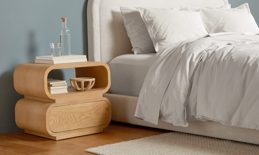 Wooden nightstand with sumptuous curves and an organic finish near a white upholstered bed.