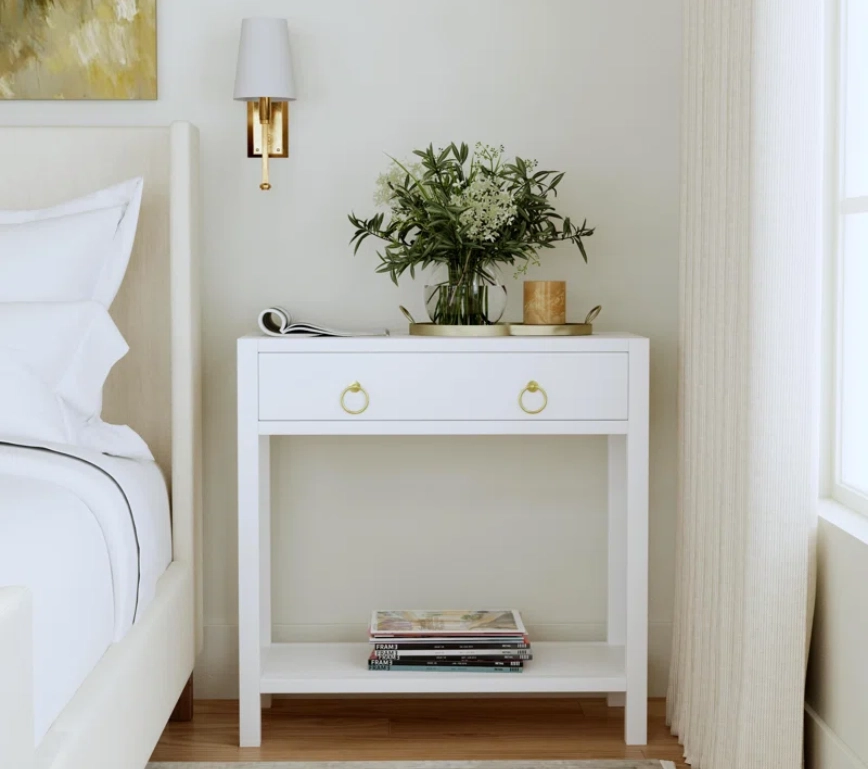 white nightstand with a shelf and one drawer with a plant on the top, on one side of the bed