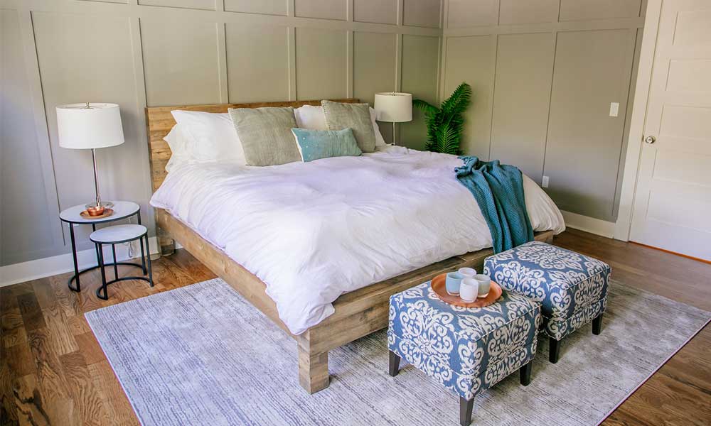 Bedroom with a woodeedn bedframe and white bedding a light gray rug, rounded side tables