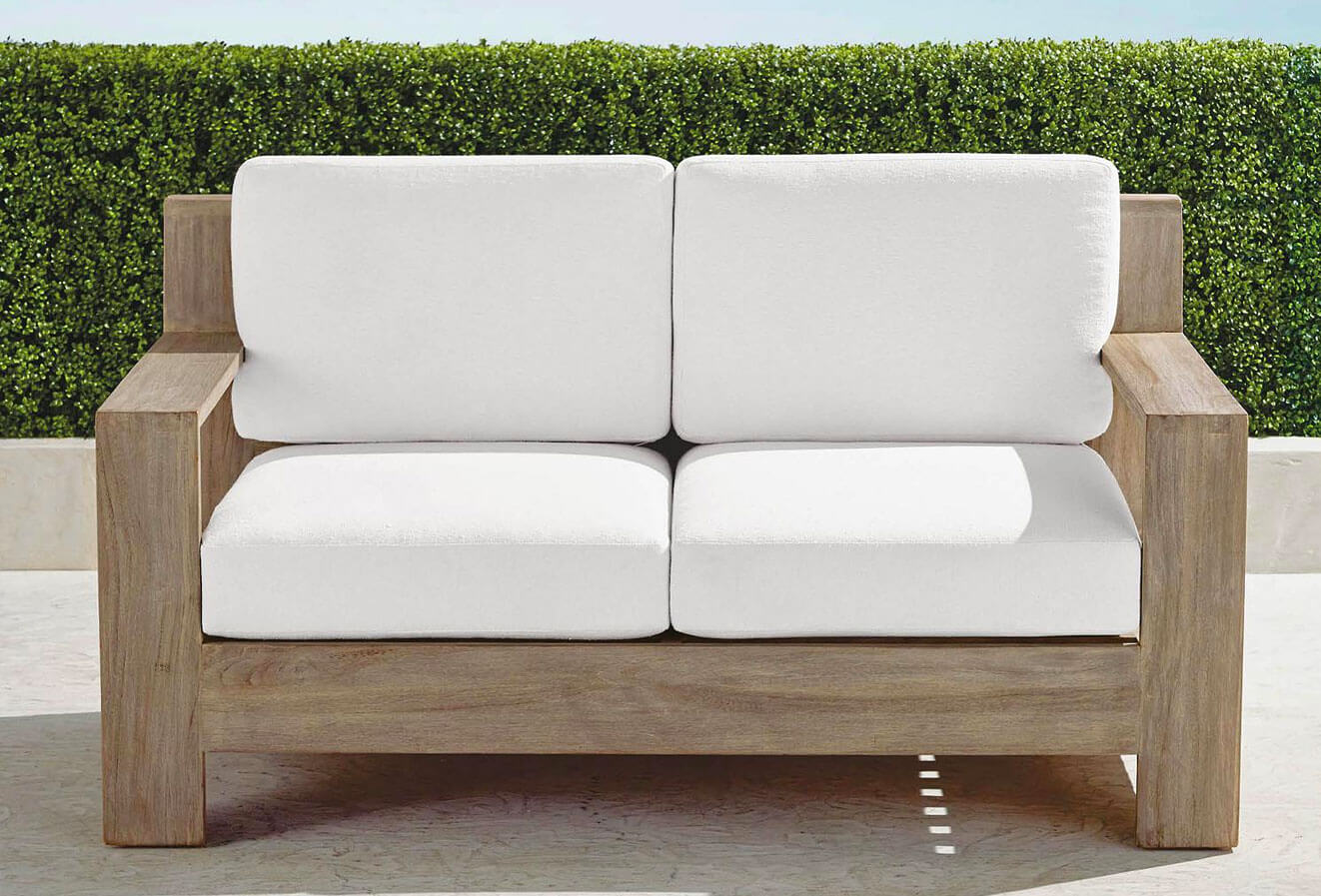 Teak loveseatl with white cushions in a patio near a green wall 