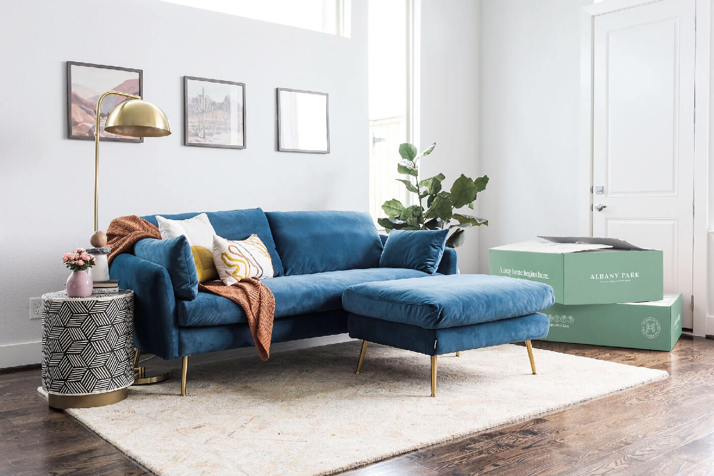 blue velvet upholstered sofa with golden legs and ottoman in a living room with white rug and boxes