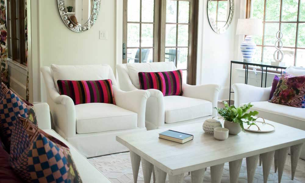 a bright living room with white furniture, colorful pillows on white chairs and a mirror