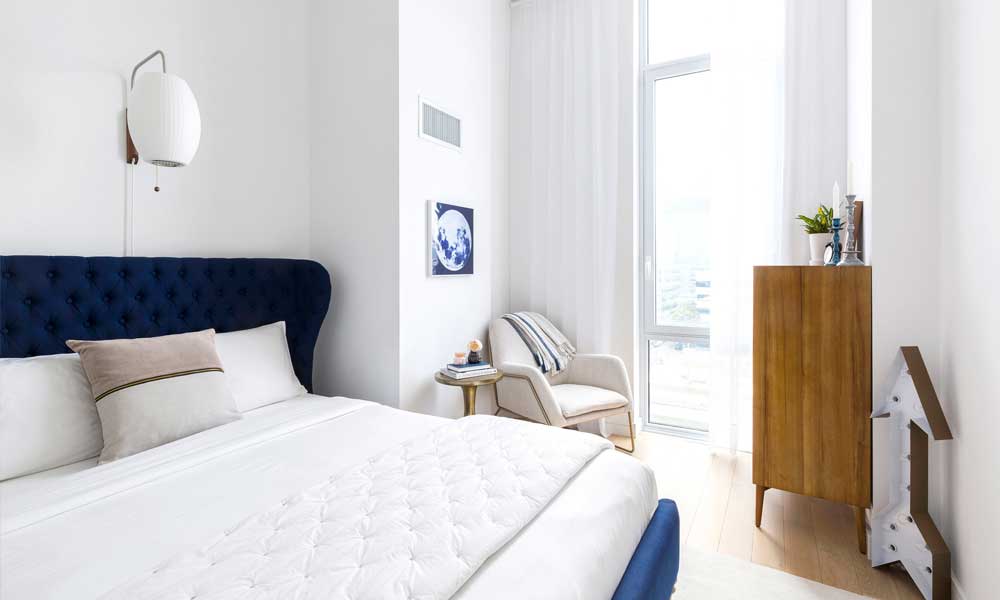 White upholstered accent char placed by the window in a bright bedroom with a blue headboard bed