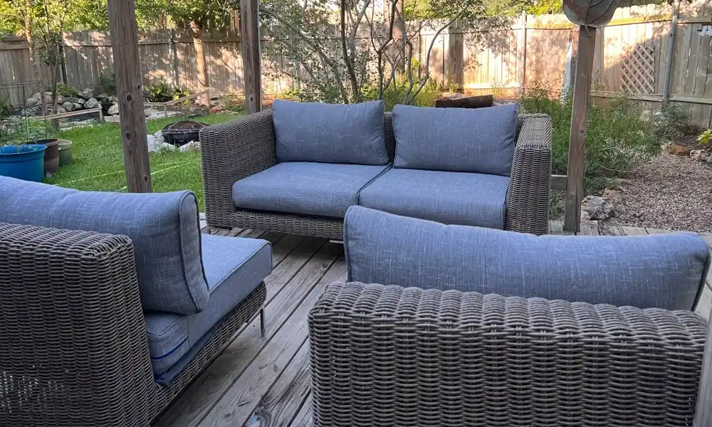 Gray wicker patio furniture with blue cushions on a wooden deck on a garden and wooden fence.