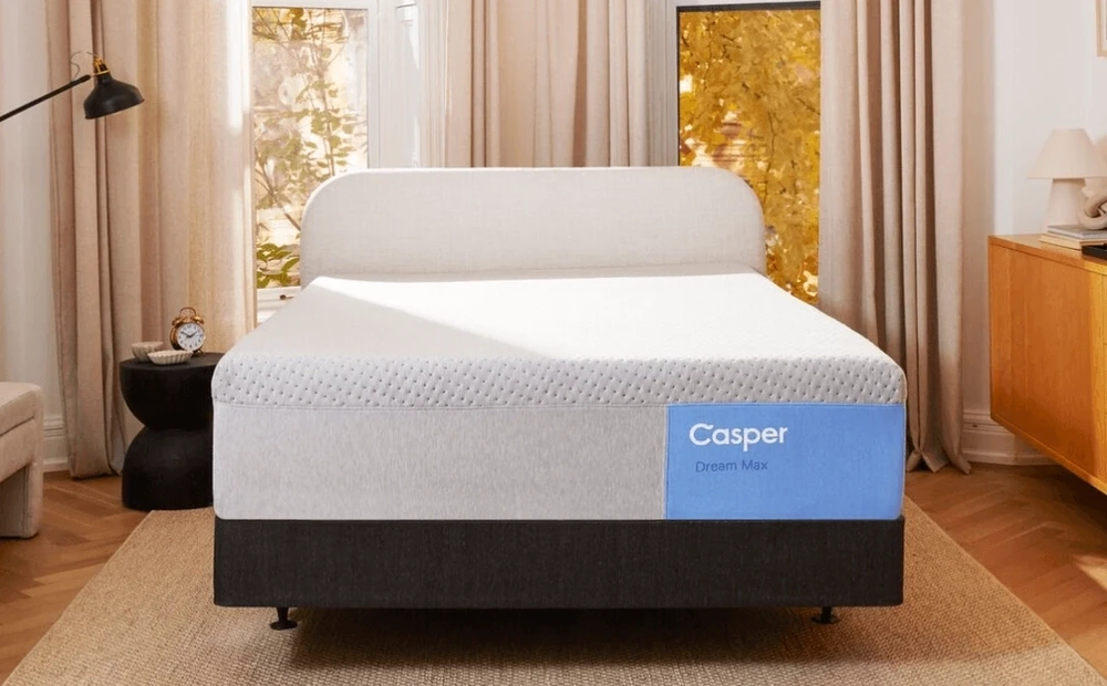 casper mattress in a bedframe with no sheets in a bright bedroom with beige curtains