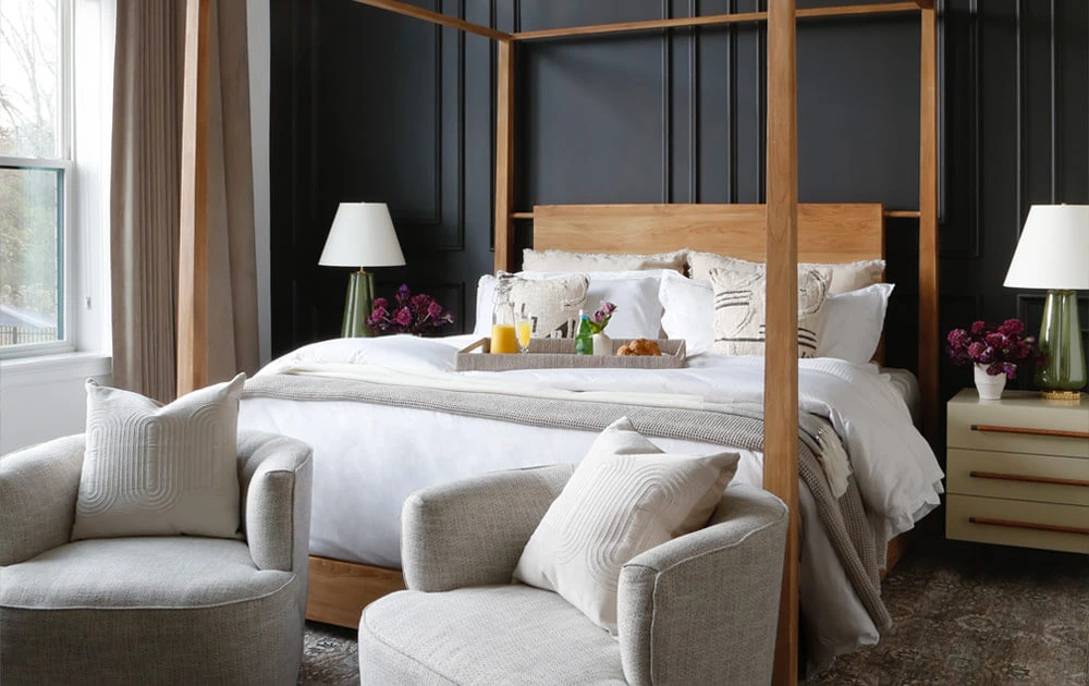 Nightstand next to a wooden canopy bed in a room with a main black wall and grey accent chairs