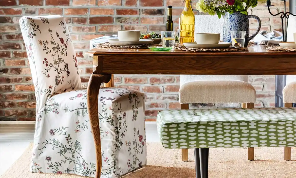 Slip-covered end chair at the end of a rectangular wooden table. A bench and crockery on the table
