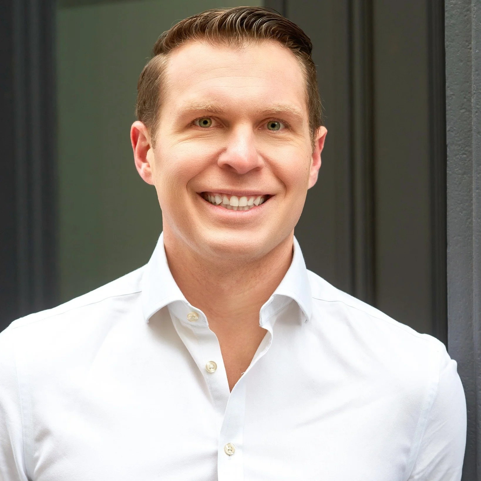 Smiling man with light brown hair wearing a white button-up shirt in front of a dark background.