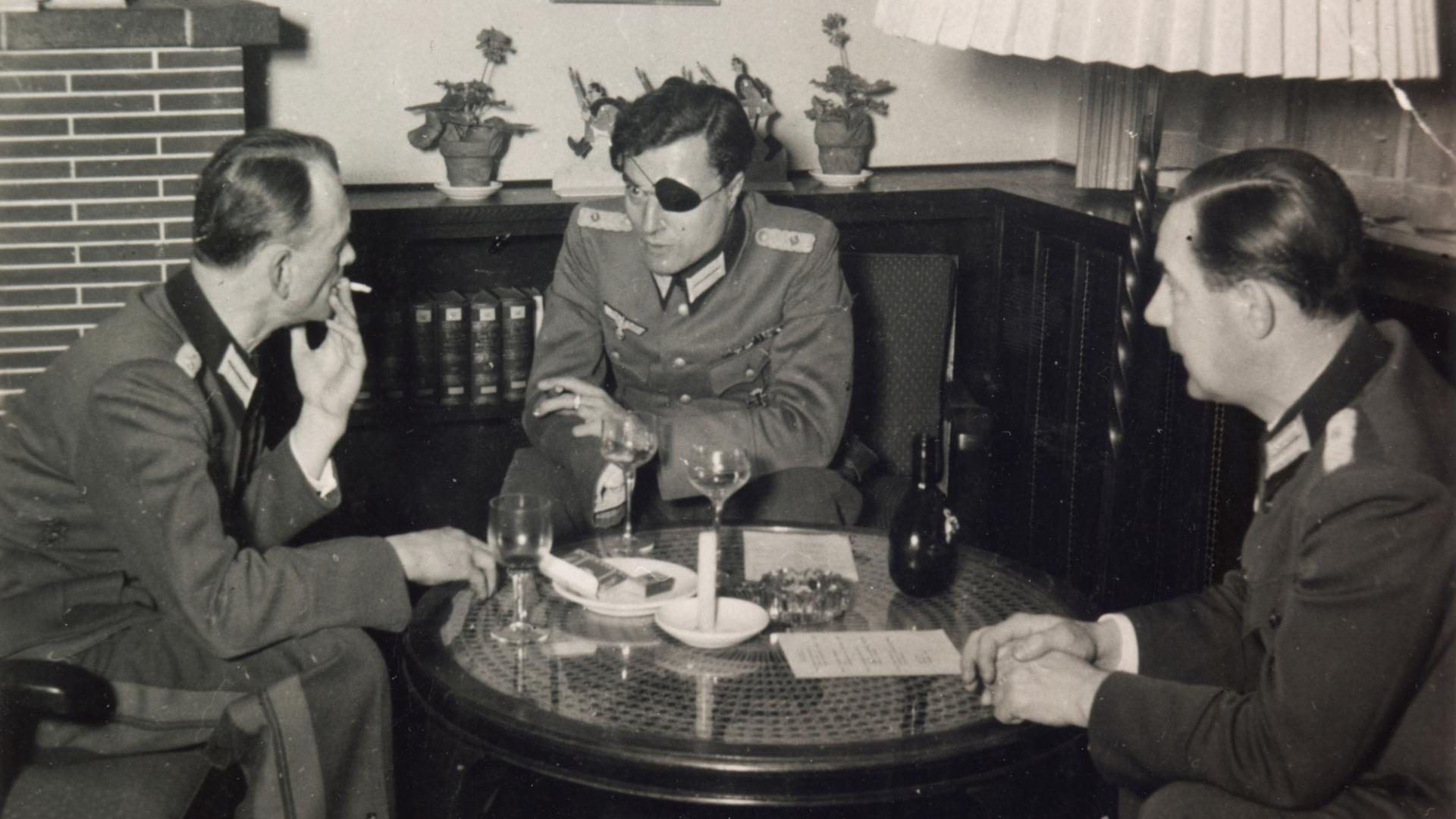 Three German army officers in uniform sit talking around a small table with wine glasses; the officer in the centre wears an eye patch