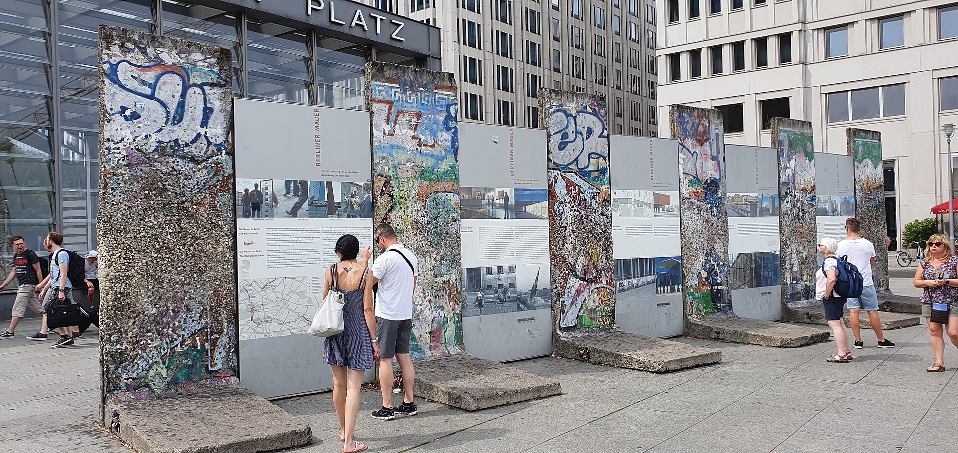 Surviving graffiti-covered segments of the Berlin Wall on display at Potsdamer Platz, with visitors reading the "Berliner Mauer" information panels between them