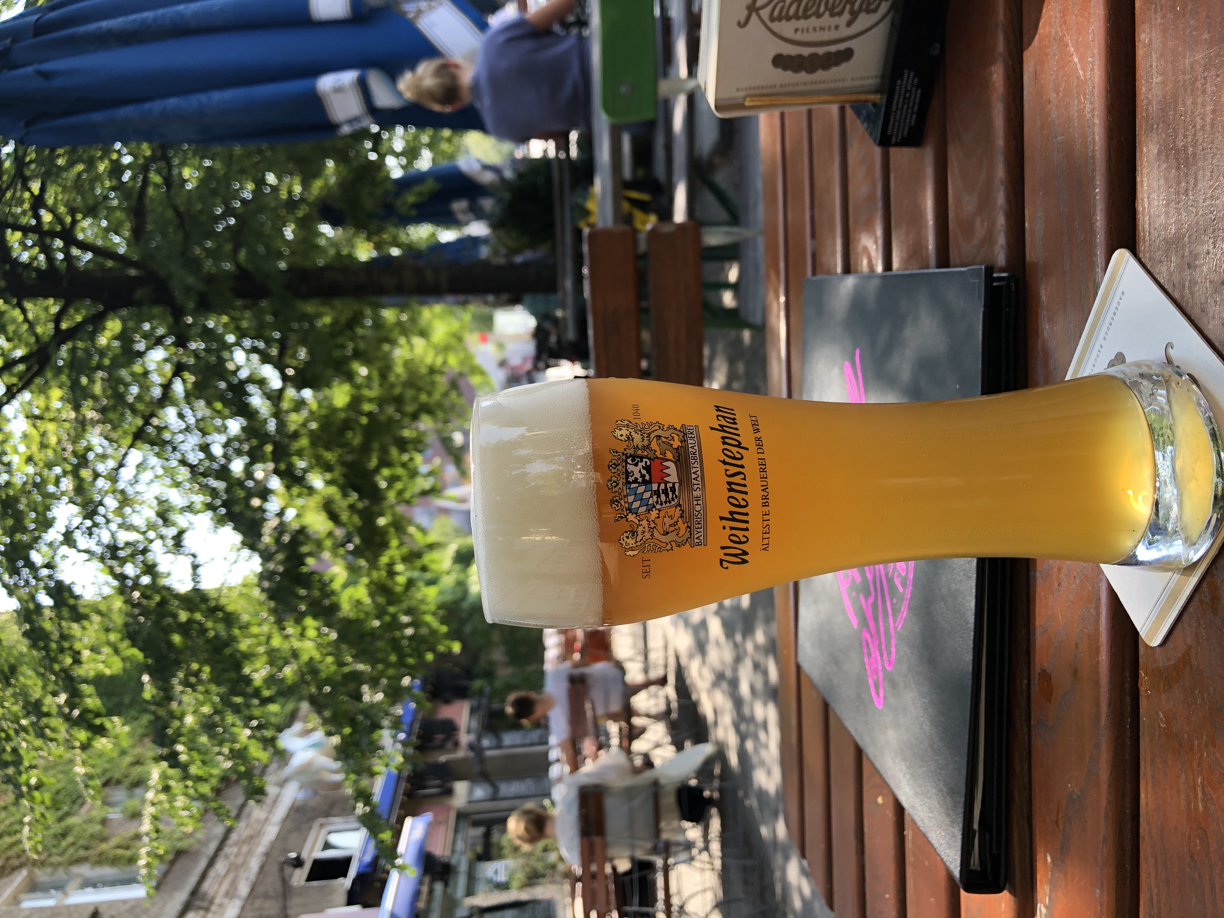 Tall glass of Weihenstephan wheat beer served in a Berlin beer garden, with outdoor seating and summer atmosphere in the background.