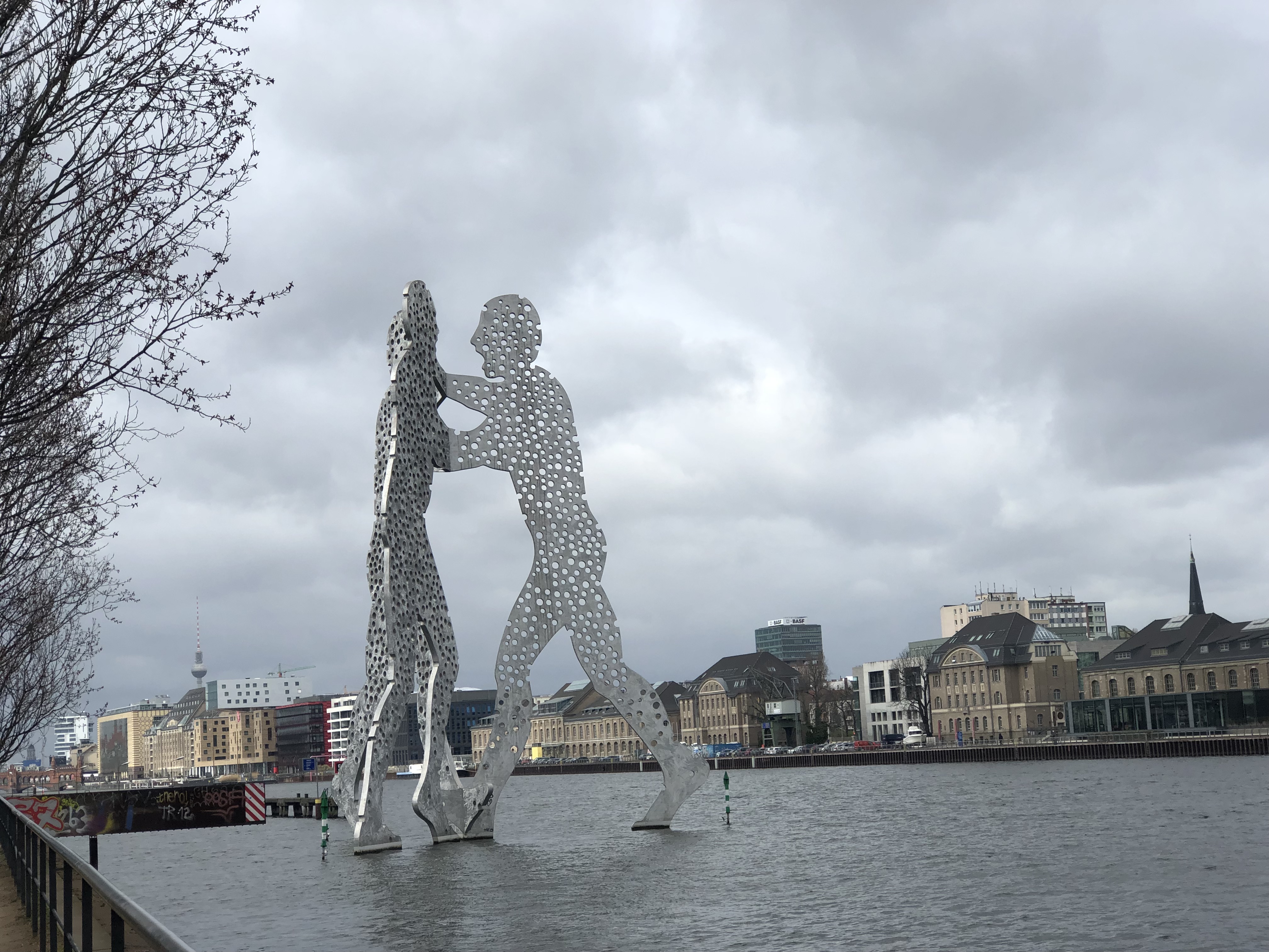 The Molecule Man sculpture on the Spree River in Berlin, with city buildings and the Fernsehturm TV Tower in the background under cloudy skies.