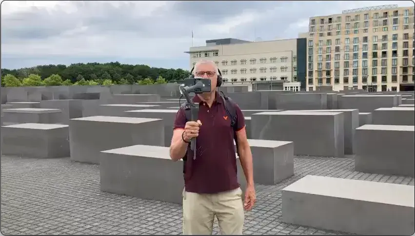 Jeremy Minsberg, also known as The Berlin Expert, is conducting a virtual tour at the Memorial to the Murdered Jews of Europe in Berlin. He is holding a gimbal-mounted smartphone while wearing headphones, ensuring a steady and immersive experience for his audience. The background features the iconic concrete stelae of the Holocaust Memorial, along with modern buildings and lush greenery under a cloudy sky. His focused expression and professional setup highlight his expertise in historical tours.