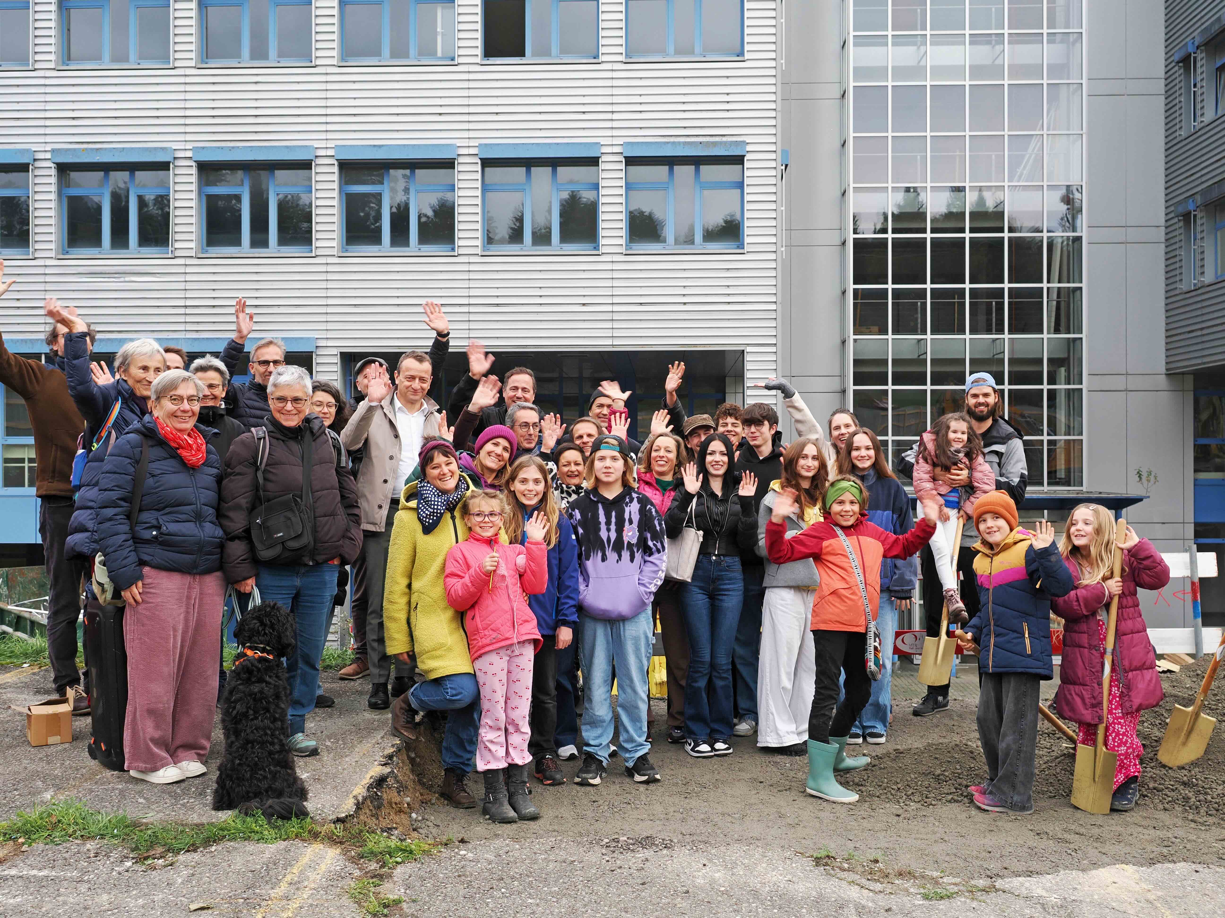 Eine Gruppe von Erwachsenen und Kindern winkt vor dem ehemaligen Verwaltungsgebäude des Weberguts in Zollikofen in die Kamera.