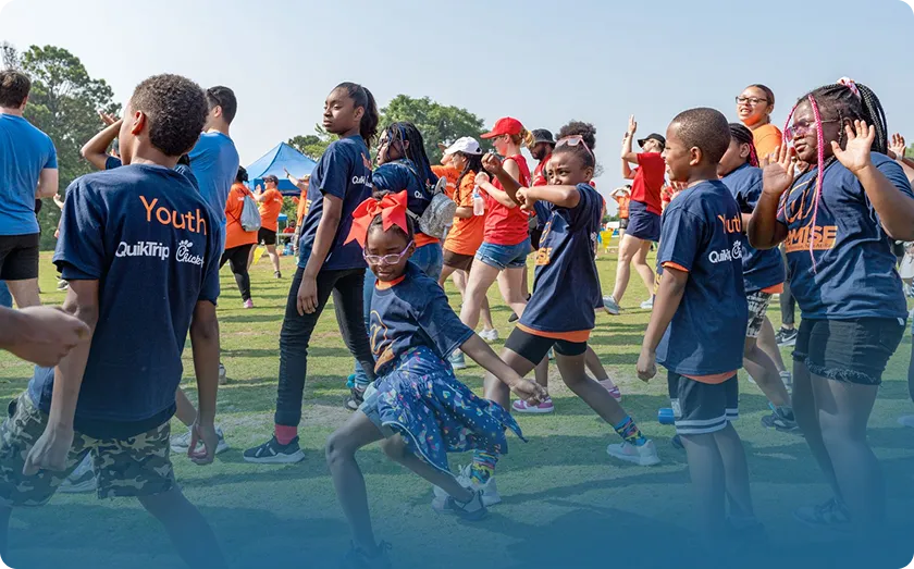 Group of children wearing navy blue Youth QuikTrip t-shirts participating in an outdoor activity on grass with adults in the background.