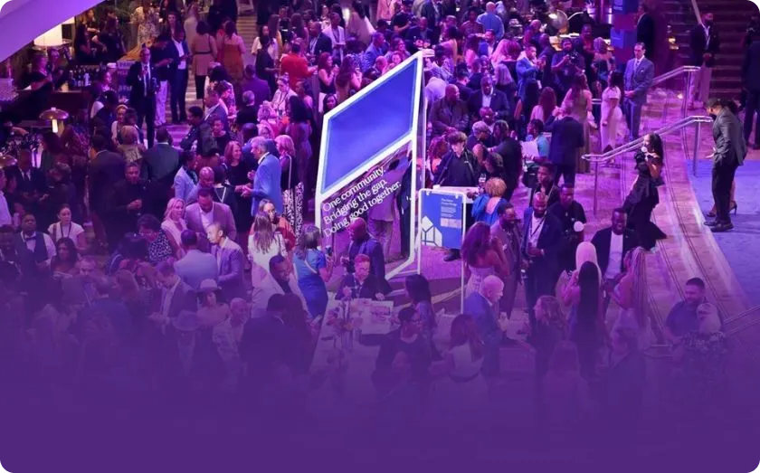 Crowded indoor event with people mingling around billboard signs and a woman speaking on stage.