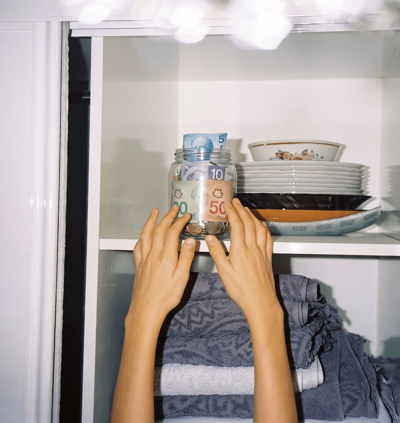 Hands reaching for a jar filled with Canadian money on a shelf with dishes and folded towels.
