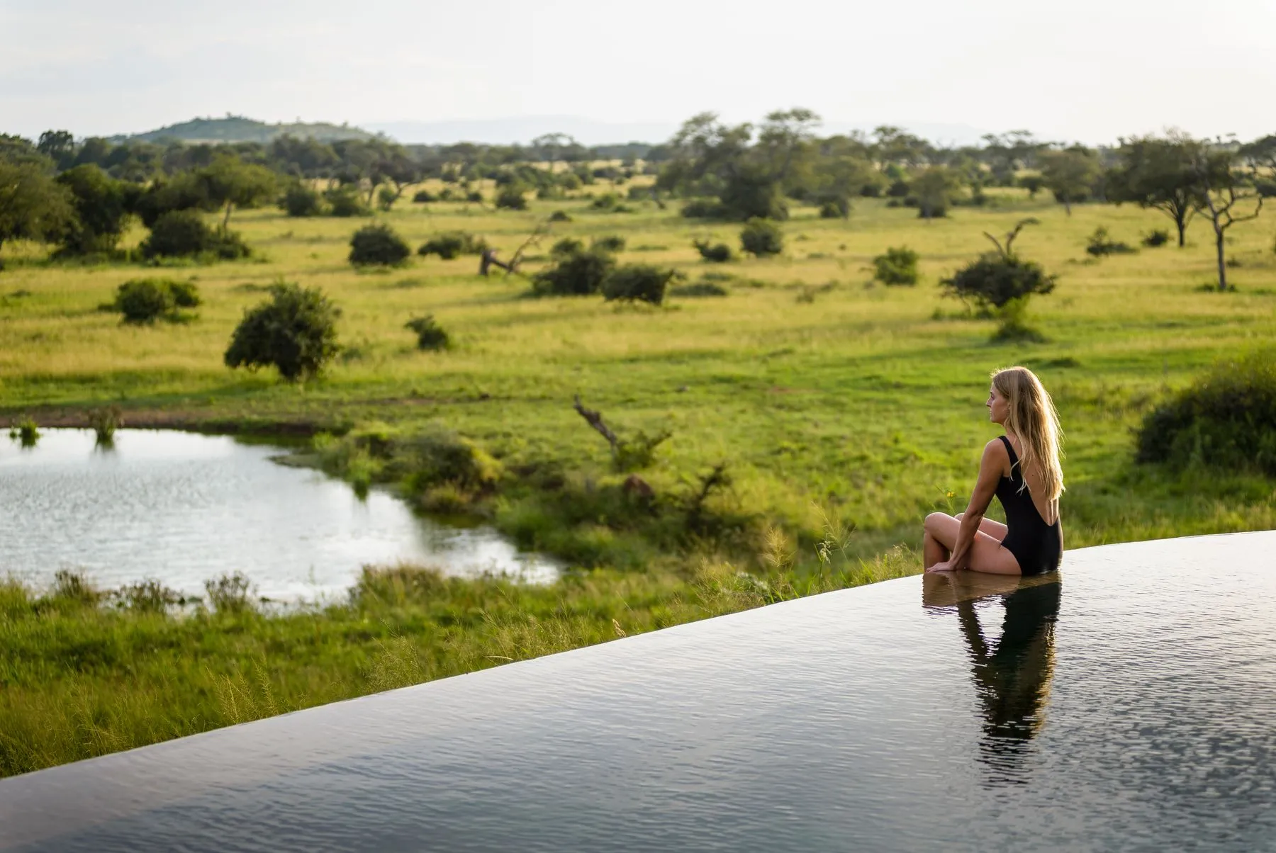 Woman sitting on the edge of a rimflow pool overlooking a serengeti landscape with green grass and acacia trees. Singita Faru Faru