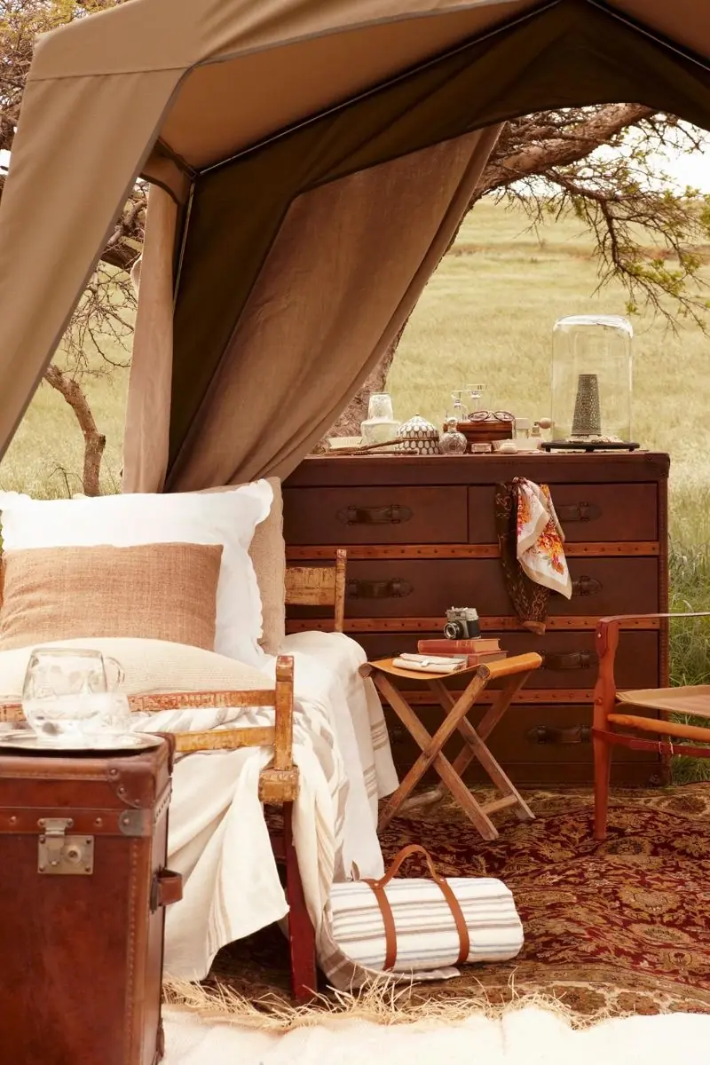 Canvas tent opening with a campaign chest of drawers, leather trunk, folding stool and patterned rug on open plains