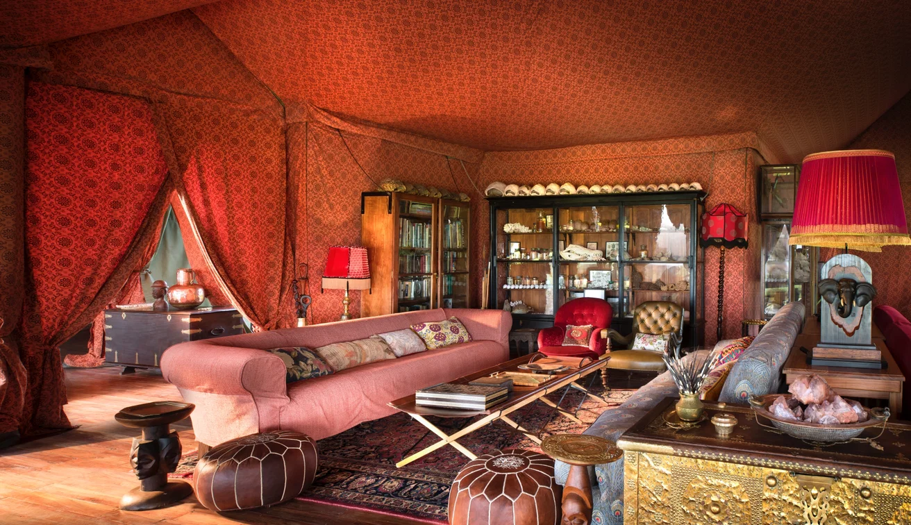 Red patterned canvas mess tent lounge with sofas, rugs and natural history cabinets at Jack’s Camp, Botswana