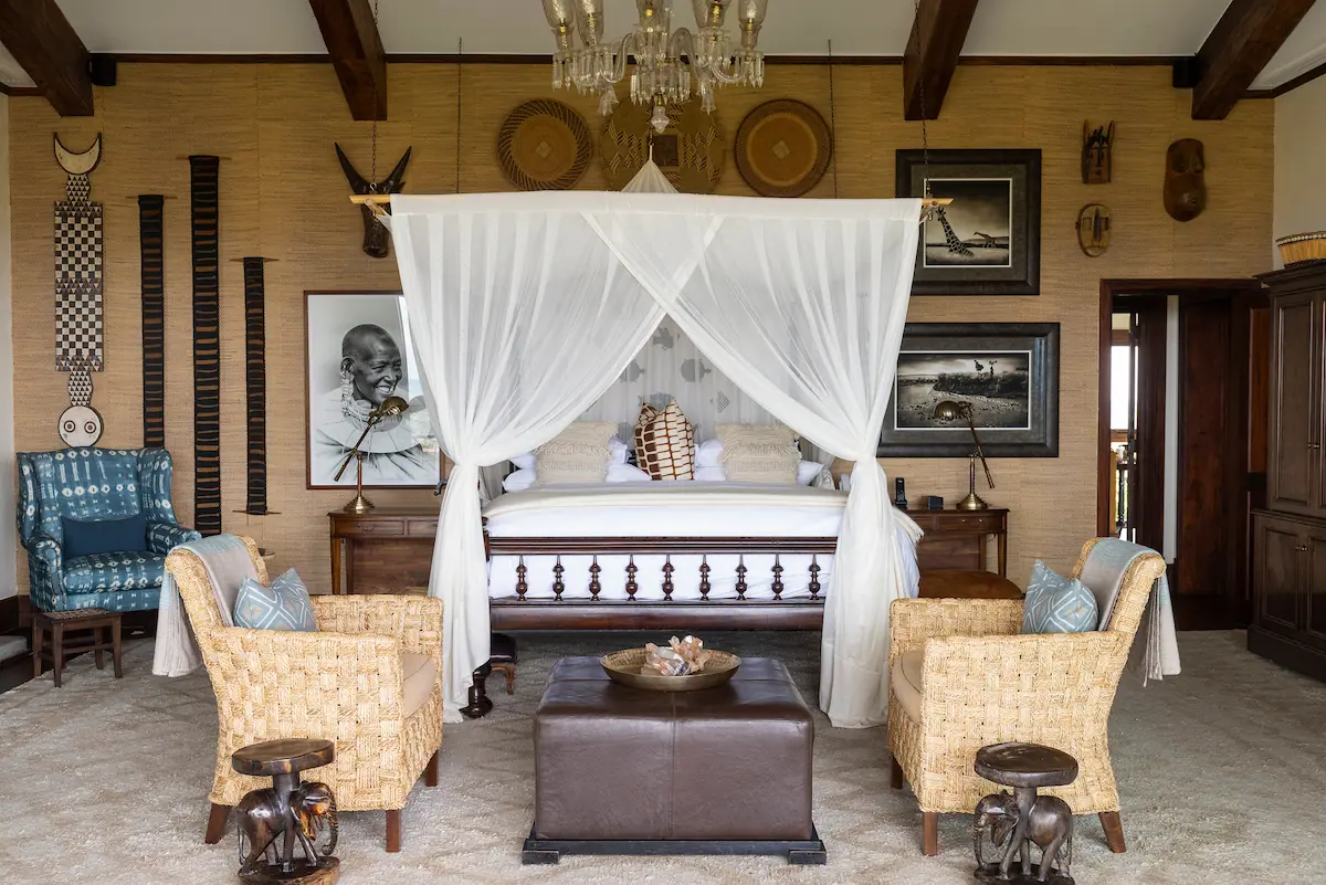 Four-poster bed with white canopy, wicker armchairs and framed photographs in a Singita Sasakwa Lodge suite, Tanzania