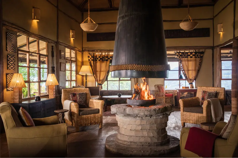 Central stone fireplace with a black hood, wicker armchairs and mountain views in the main lounge at Virunga Lodge, Rwanda