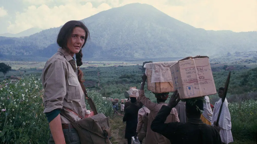 Archival photograph of Dian Fossey on a mountain trail, porters carrying boxes and a volcano behind