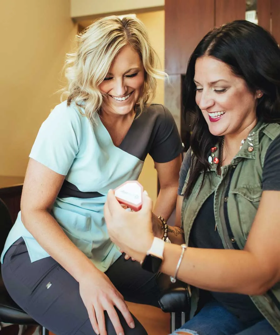 Two women smiling and examining a dental product together indoors. One woman is holding a small jar, showing it to the other.