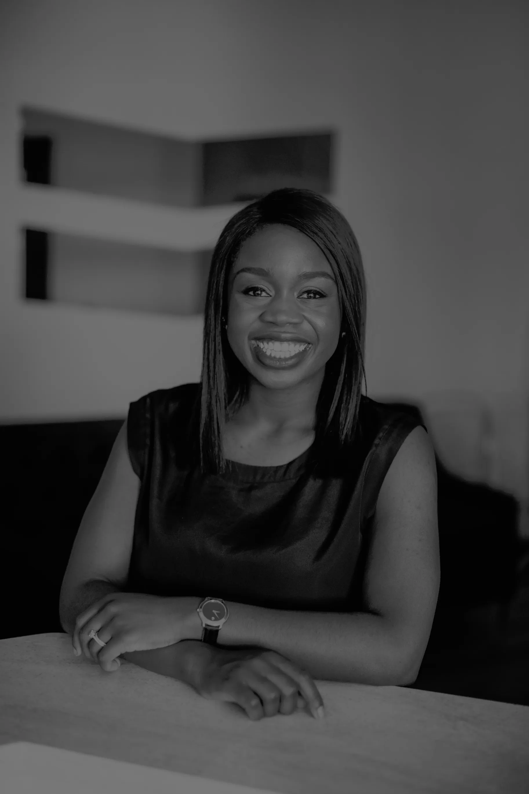 A black and white photo of a smiling dentist with straight black hair, wearing a sleeveless top and a watch, resting her arms on a table in a dimly lit room, perfect for dental marketing