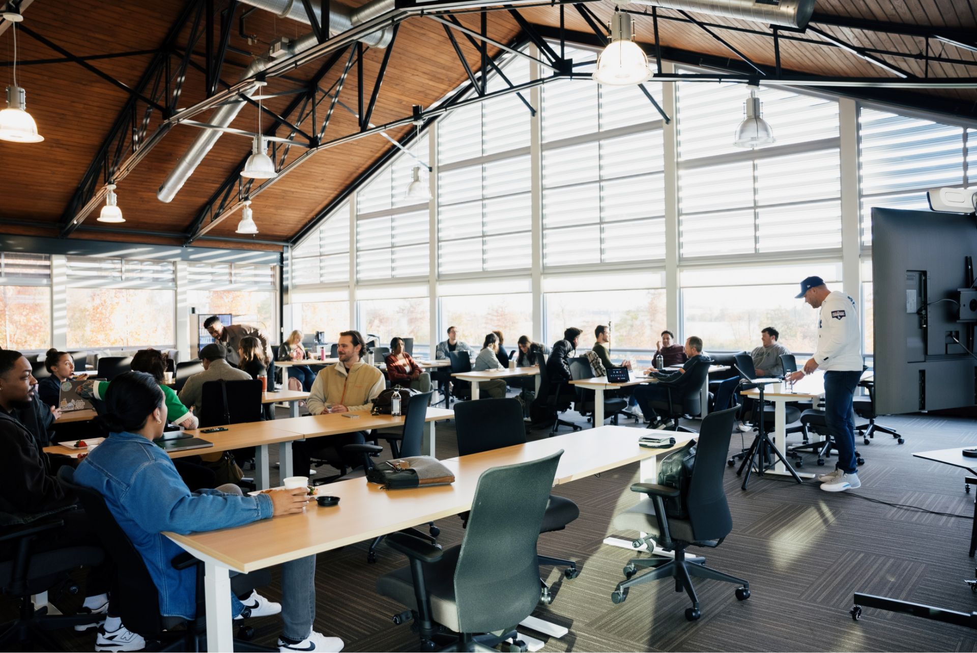 A group of people seated at desks in a modern, well-lit office space listen to a person presenting at the front of the room.