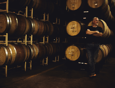 Man in dark clothing leaning against stacked wooden barrels inside a dimly lit cellar or storage room.