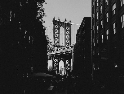 Black and white photo of the Manhattan Bridge framed by buildings with the Empire State Building visible in the distance.