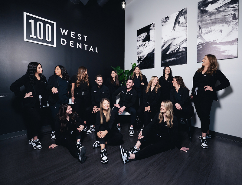 Group of dental office staff members in black uniforms posing and smiling in a modern lobby with 100 West Dental sign on the wall.