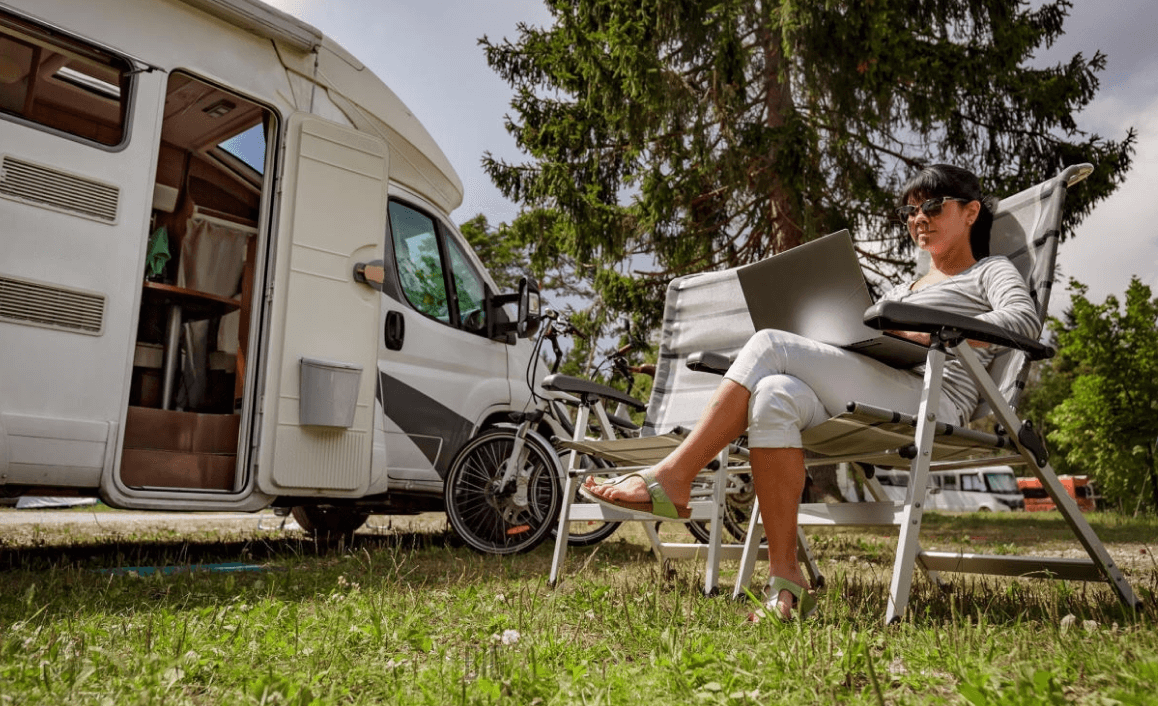 A woman uses a laptop in a folding chair next to an open RV and bicycle, enjoying remote work amidst a sunny, grassy campsite