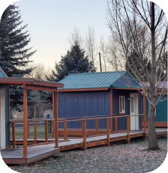 A small cabin with a blue roof, featuring a wooden walkway leading to its entrance, surrounded by nature.