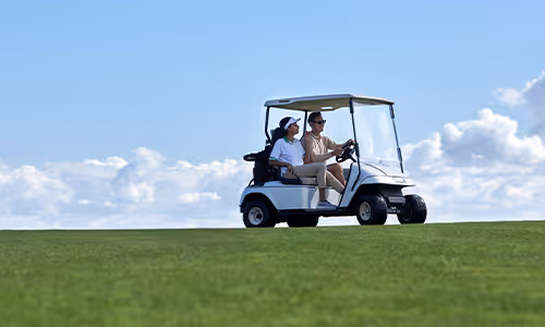 Two people seated in a white golf cart driving across a green fairway under a blue sky