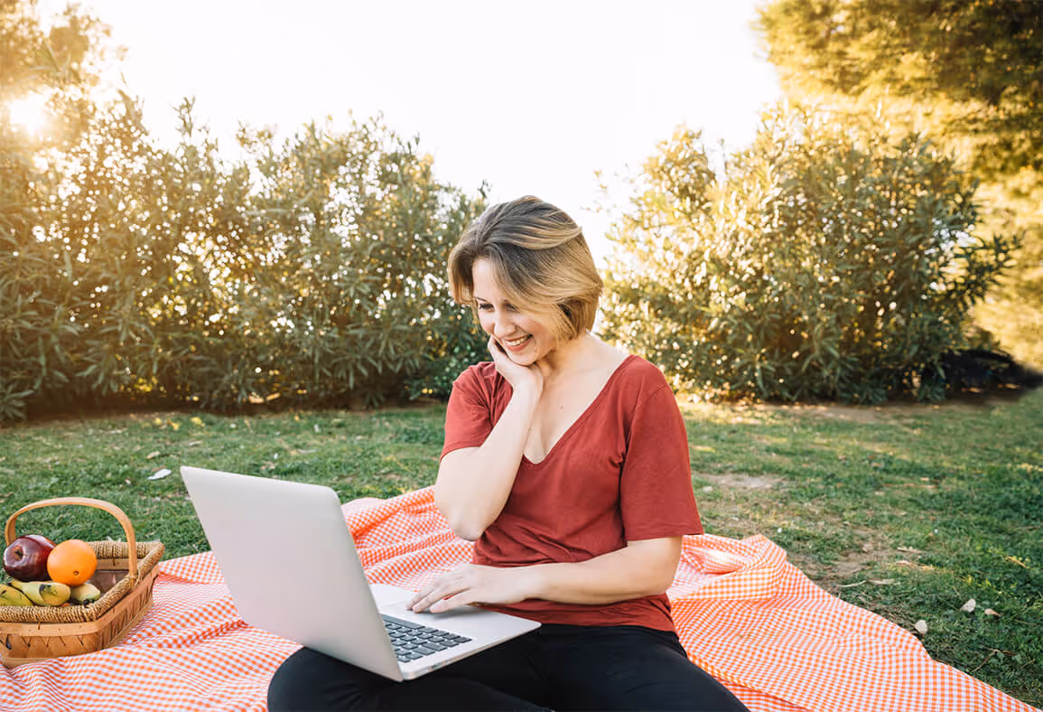 Woman seated on a picnic blanket in a park, smiling at her laptop with a basket of fruit beside her.